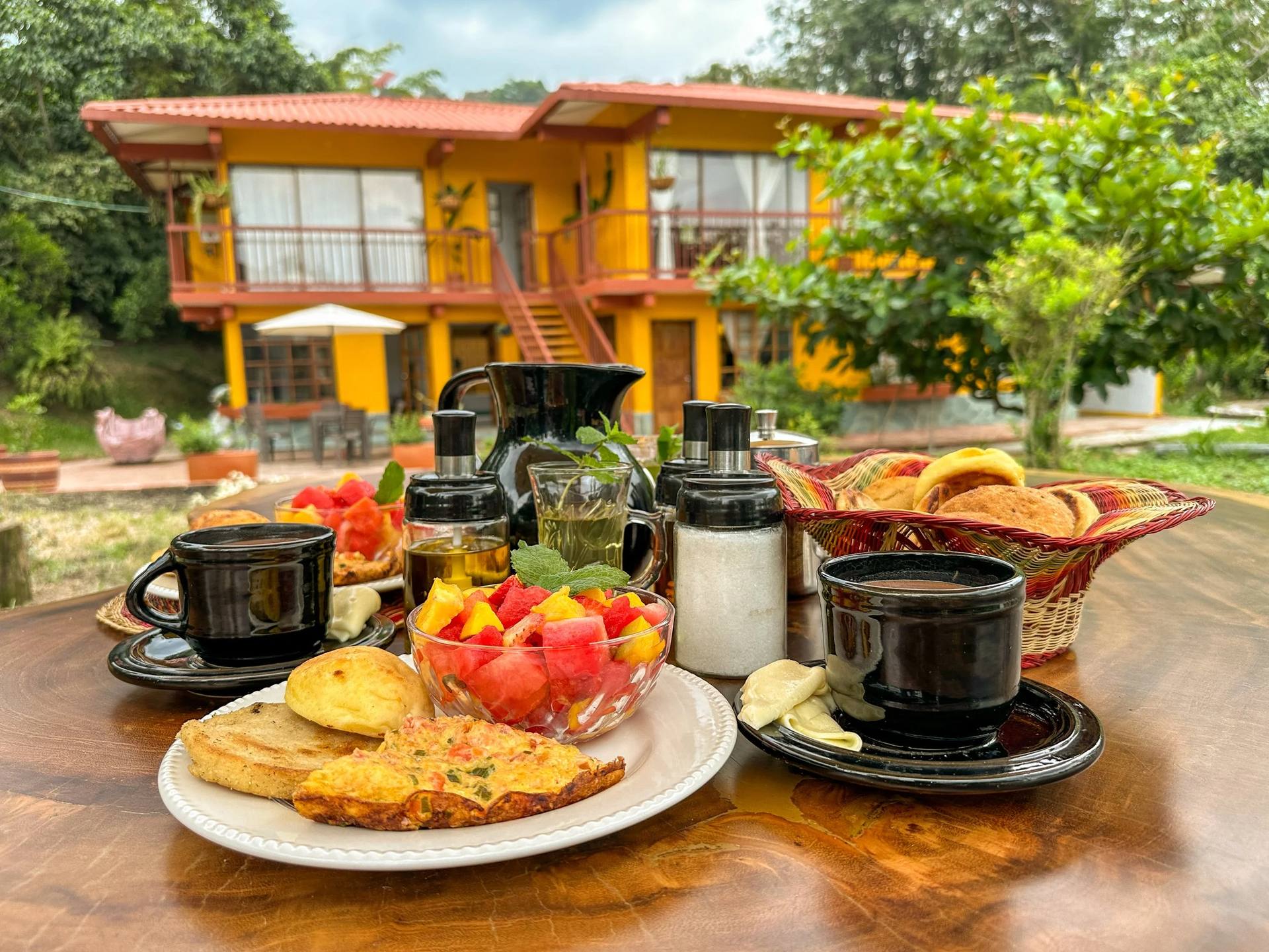 Desayuno con frutas, panes, café y otros alimentos en una mesa con una casa amarilla de fondo