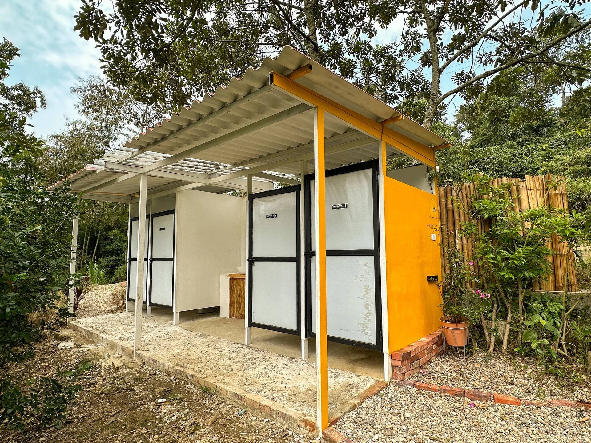 Outdoor bathroom block with orange and white walls and black doors, under a white metal roof, surrounded by greenery